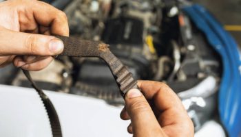 An auto mechanic shows a torn timing belt with worn teeth against the background of an open car hood close-up